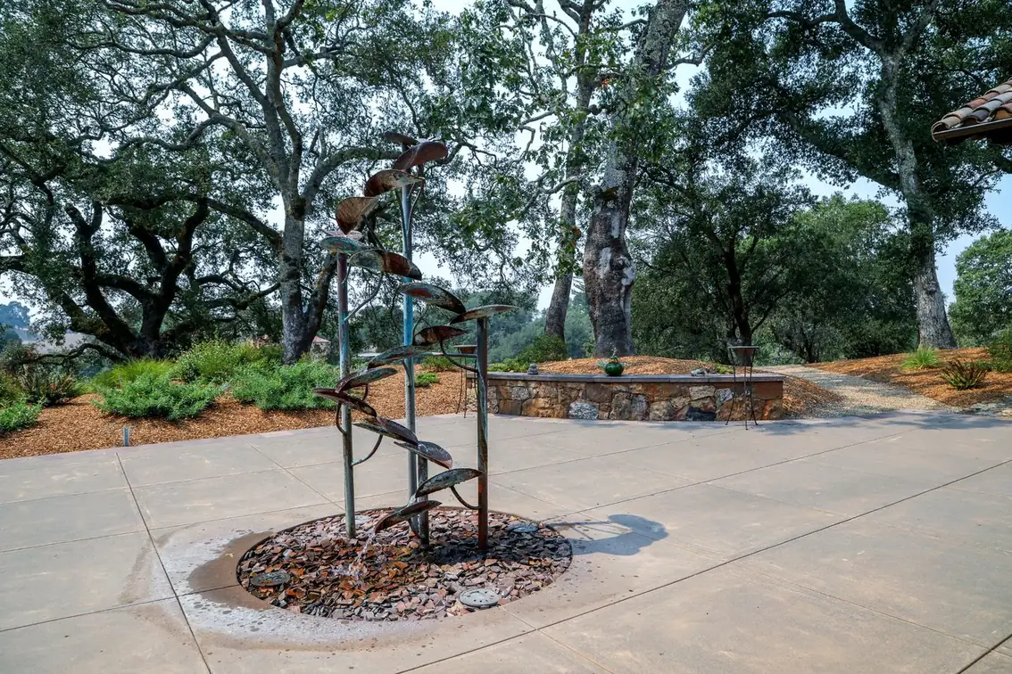 A copper leaf fountain on a concrete patio beneath mature oaks, with a stone wall behind