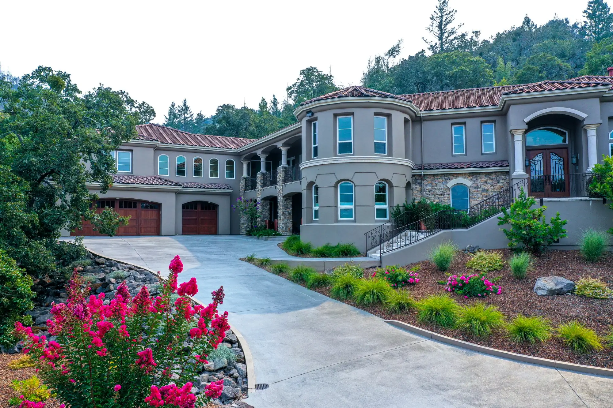 A stucco and tile-roofed estate with a curved driveway, crepe myrtle in bloom, and ornamental grasses