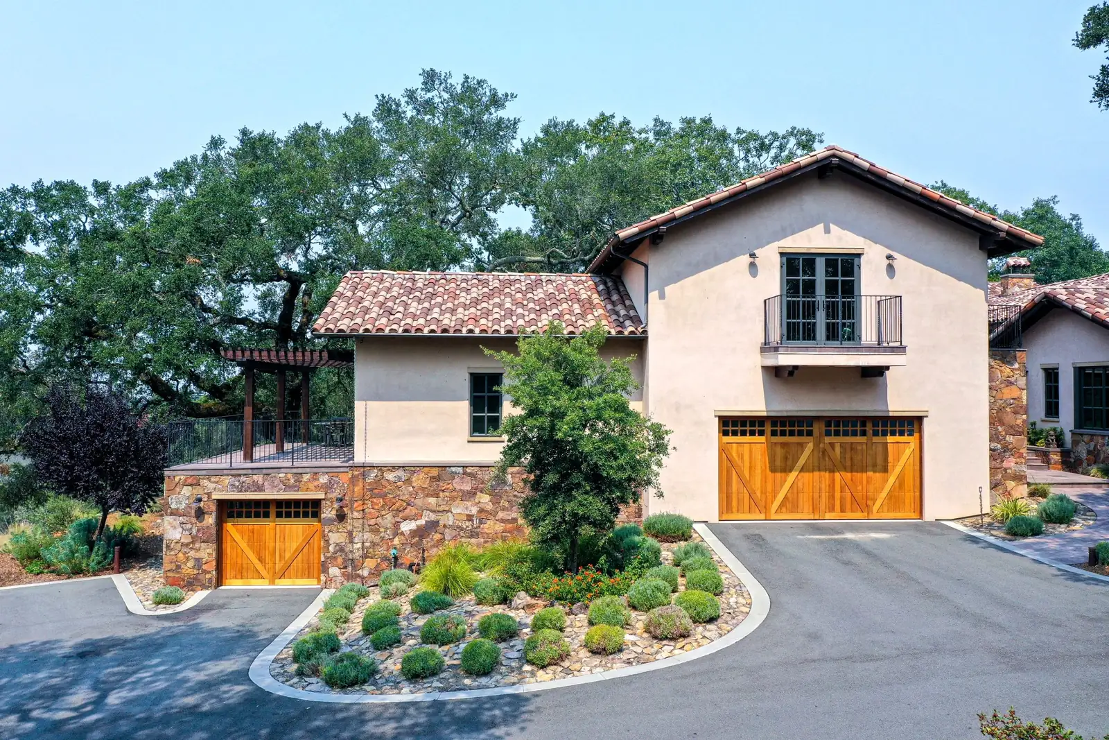 Stucco home with wooden garage doors, a river-rock bed of mounded lavender and a large oak behind